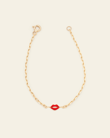 A photo of a Solid Yellow Gold thin staple chain bracelet with a red enamel lip charm in the centre against a cream background. 