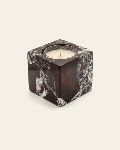 A photo of a candle set in a square, marbled dark stone holder with a smooth surface against a cream background. The holder has a unique white and dark brown marbling pattern, giving it a luxurious, artistic look. The candle features a single wick and is framed within the stone's circular top opening.