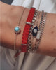 A close up video of a model’s wrist wearing six bracelets from left to right: a delicate bracelet featuring a turquoise stone encircled by white sapphires, a red woven thread bracelet with three Yellow Gold tabs with 5 diamonds on one tab, a thick curb chain bracelet, an evil eye chain bracelet with a blue enamel surface with centred clustered diamonds, a staple mix tennis chain bracelet with a centred baguette diamond tab and a delicate curb chain bracelet.