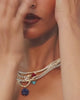 A close up video of a model wearing multiple Gold Vermeil tennis necklaces with different semiprecious stone pendants on them such as turquoise, lapis, coral and howlite and baroque pearls against a cream background. She is seen moving her hands around the necklaces showing the intricate design and draping around her neck.