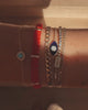 A close up video of a model’s wrist wearing six bracelets from left to right: a delicate bracelet featuring a turquoise stone encircled by white sapphires, a red woven thread bracelet with three Yellow Gold tabs with 5 diamonds on one tab, a thick curb chain bracelet, an evil eye chain bracelet with a blue enamel surface with centred clustered diamonds, a staple mix tennis chain bracelet with a centred baguette diamond tab and a delicate curb chain bracelet.